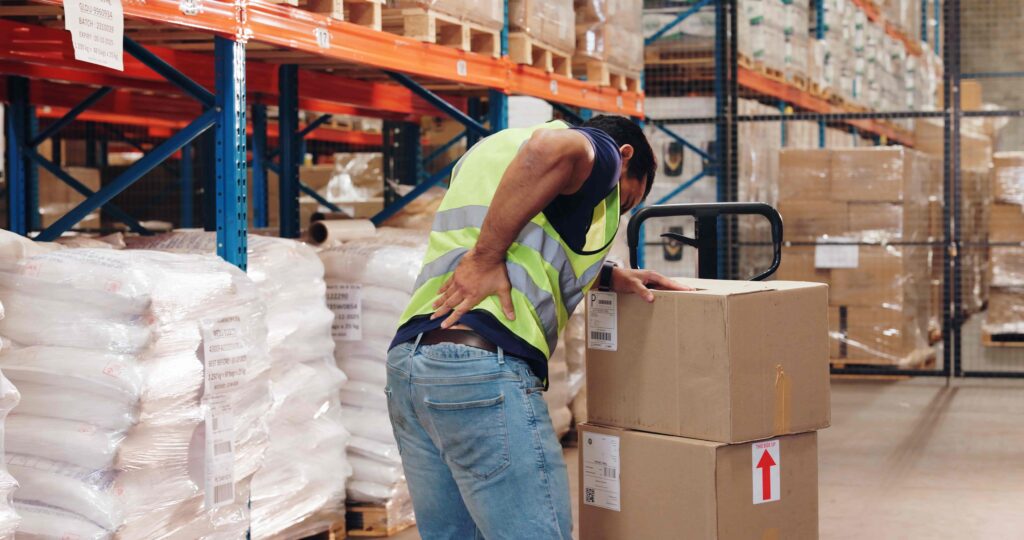 Warehouse worker in safety vest suffering back injury while lifting heavy boxes Warehouse worker in safety vest suffering back injury while lifting heavy boxes
